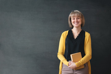Smiling teacher with textbook in hands on black blackboard background with copy space.
