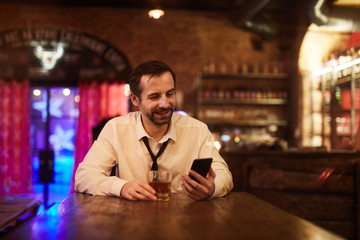 Portrait of mature businessman drinking alcohol in bar after work and smiling looking at smartphone screen, copy space