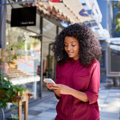 Smiling young woman standing on a sidewalk reading text messages