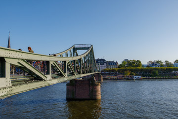 Outdoor sunny view of Eiserner Steg, historical pedestrian Iron Bridge, and promenade on riverside of Main River in sunny day in Frankfurt, Germany.