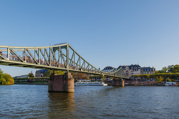 Outdoor sunny view of Eiserner Steg, historical pedestrian Iron Bridge, and promenade on riverside of Main River in sunny day in Frankfurt, Germany.