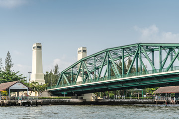 Bangkok city, Thailand - March 17, 2019: Green metal Memorial bridge over Chao Phraya River, with its two white towers under light blue sky. Some green foliage.
