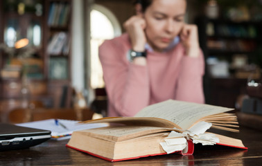 Repetition. A thick open book with many bookmarks, lies on a table. Brunette girl covering her ears with her fingers.