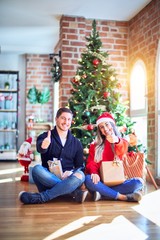 Young couple wearing santa claus hat sitting on the floor around christmas tree at home doing happy thumbs up gesture with hand. Approving expression looking at the camera with showing success.