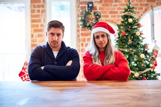 Young couple wearing santa claus hat sitting on chair and table around christmas tree at home skeptic and nervous, disapproving expression on face with crossed arms. Negative person.