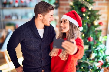 Young beautiful couple smiling happy and confident. Standing and hugging make selfie by camera around christmas tree at home