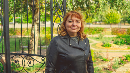 Woman against fence in garden. Content woman looking dreamily away while standing in green park against ornamental iron fence