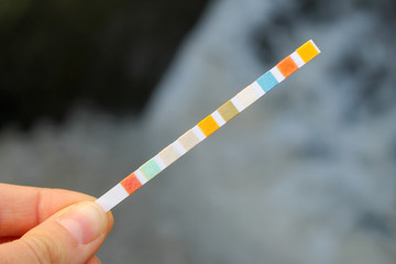 Strip for water quality measurement held in fingers (female hand). Ten multicolor squares for chemical testing of different reagents in water to identify its purity. Background of waterfall. - obrazy, fototapety, plakaty