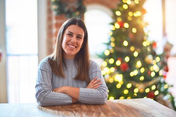 Young beautiful woman sitting at the table at home around christmas tree and decoration happy face smiling with crossed arms looking at the camera. Positive person.