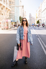 Young woman in an pink dress, denim jacket, boots and eye cat sunglasses standing at the famous view of Gran Via main broadway road in the downtown of Madrid, Spain .