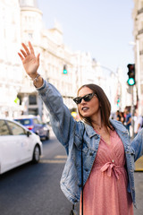 Young brunette woman asking for a taxi raising her arms in Madrid city. Fashion model wearing pink dress, denim jacket, boots and eye cat sunglasses. Female calling a taxi. Transportation concept.
