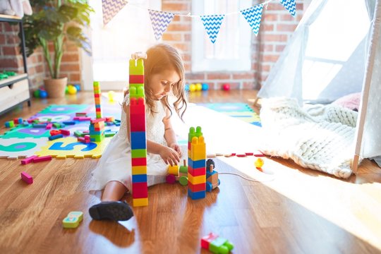 Adorable blonde toddler playing with building blocks around lots of toys at kindergarten