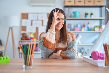 Young beautiful teacher woman wearing sweater and glasses sitting on desk at kindergarten doing ok gesture with hand smiling, eye looking through fingers with happy face.