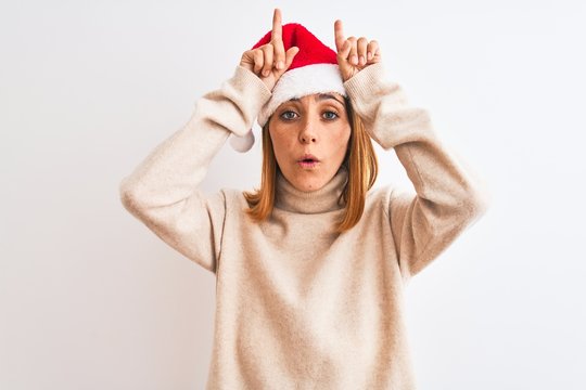 Beautiful redhead woman wearing christmas hat over isolated background doing funny gesture with finger over head as bull horns