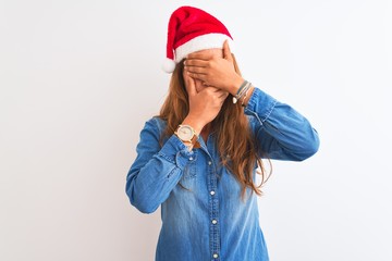 Young beautiful redhead woman wearing christmas hat over isolated background Covering eyes and mouth with hands, surprised and shocked. Hiding emotion