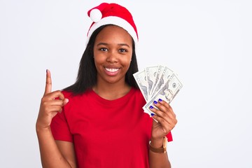 Young african american woman wearing christmas hat and holding bunch of dollars banknotes surprised with an idea or question pointing finger with happy face, number one