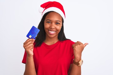 Young african american woman wearing christmas hat and holding credit card pointing and showing with thumb up to the side with happy face smiling