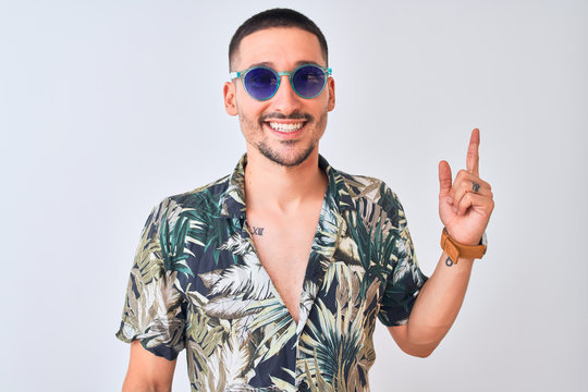 Young handsome man wearing Hawaiian sumer shirt and blue sunglasses over isolated background with a big smile on face, pointing with hand and finger to the side looking at the camera.