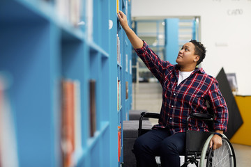 Portrait of disabled student in wheelchair choosing books while studying in college library, copy...