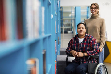 Portrait of two college students, one of them in wheelchair, posing together standing by shelves in library, copy space