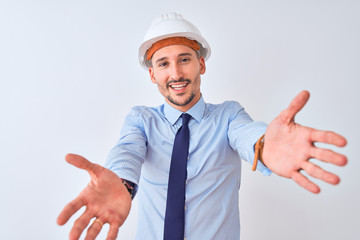 Young business man wearing contractor safety helmet over isolated background looking at the camera smiling with open arms for hug. Cheerful expression embracing happiness.