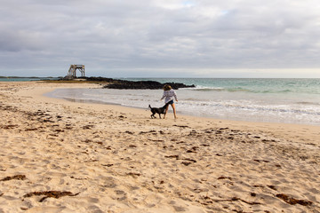 Woman and dog romping on beautiful sand beach with wooden platform lookout in the background, Puerto Villamil, Isabela Island, Galapagos, Ecuador