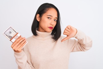 Young beautiful chinese woman holding dollars standing over isolated white background with angry...