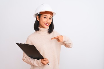 Chinese architect woman wearing helmet holding clipboard over isolated white background with surprise face pointing finger to himself