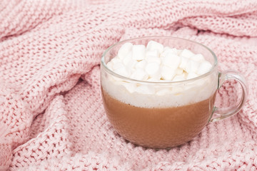 Transparent cup of cocoa with marshmallow and gift box with bow on pink knitted plaid, close up. Winter cozy concept
