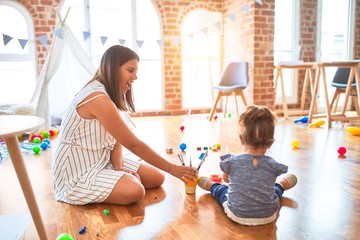 Young beautiful teacher and toddler sitting on the floor playing with brush at kindergarten