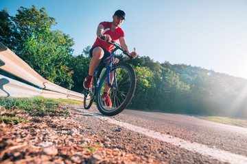 Fototapeta premium Fit male biker cyclist riding his bike cycle on an asphalt road at sunset while the sun sets through his wheel.