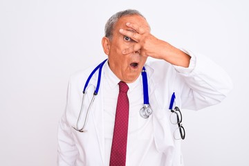 Senior grey-haired doctor man wearing stethoscope standing over isolated white background peeking in shock covering face and eyes with hand, looking through fingers with embarrassed expression.