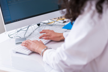 Close up hands of a female physician ( nurse ) typing on her white keyboard while sitting on a white desk in a hospital lobby.