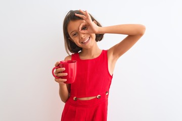 Obraz na płótnie Canvas Beautiful child girl holding red cup of tea standing over isolated white background with happy face smiling doing ok sign with hand on eye looking through fingers