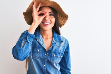 Young beautiful woman wearing denim shirt and hat standing over isolated white background doing ok gesture with hand smiling, eye looking through fingers with happy face.