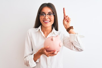 Beautiful businesswoman wearing glasses holding piggy bank over isolated white background surprised with an idea or question pointing finger with happy face, number one