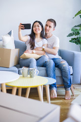 Young beautiful couple sitting on the sofa using smartphone to take a selfie at new home around cardboard boxes