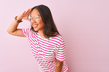 Young chinese woman wearing striped t-shirt and glasses over isolated pink background very happy and smiling looking far away with hand over head. Searching concept.