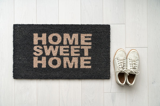New home moving in door mat entrance welcome doormat with text writing HOME SWEET HOME and white sneakers of happy condo homeowner. Top view of rug and wooden floor.