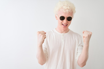 Young albino blond man wearing t-shirt and sunglasses over isolated white background celebrating surprised and amazed for success with arms raised and open eyes. Winner concept.