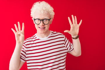 Young albino blond man wearing striped t-shirt and glasses over isolated red background showing and pointing up with fingers number nine while smiling confident and happy.