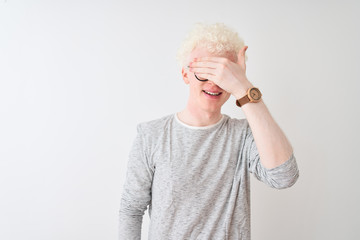 Young albino blond man wearing striped t-shirt and glasses over isolated white background smiling and laughing with hand on face covering eyes for surprise. Blind concept.