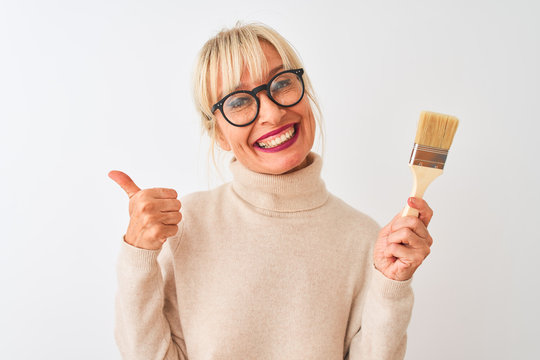 Middle age painter woman holding paint brush standing over isolated white background happy with big smile doing ok sign, thumb up with fingers, excellent sign