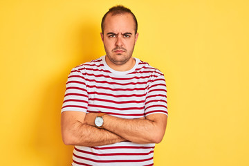 Young man wearing casual striped t-shirt standing over isolated yellow background skeptic and nervous, disapproving expression on face with crossed arms. Negative person.