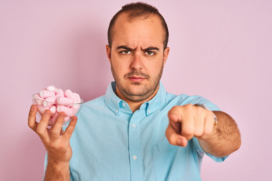 Young man holding bowl with marshmallows standing over isolated pink background pointing with finger to the camera and to you, hand sign, positive and confident gesture from the front