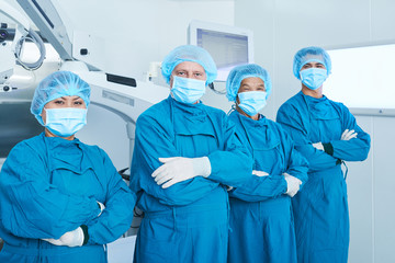 Confident positive surgical team in uniform standing in operating room with their arms folded and looking at camera
