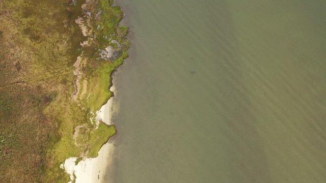 top down drone camera view of the shoreline in a marsh area, on a cloudy day. Green waters in the channel below, with a drastic contrast against the beige sand & brownish, green grass