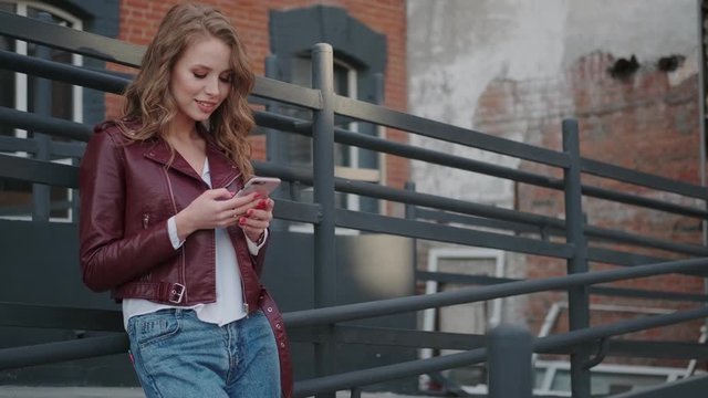 beautiful young woman uses a smartphone while standing against the backdrop of urban buildings