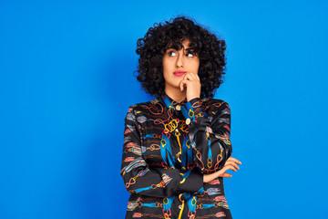 Young arab woman with curly hair wearing colorful shirt over isolated blue background with hand on chin thinking about question, pensive expression. Smiling with thoughtful face. Doubt concept.