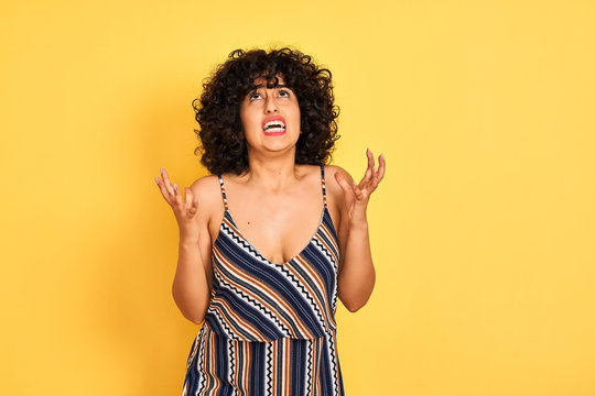 Arab woman with curly hair wearing striped colorful dress over isolated yellow background crazy and mad shouting and yelling with aggressive expression and arms raised. Frustration concept.